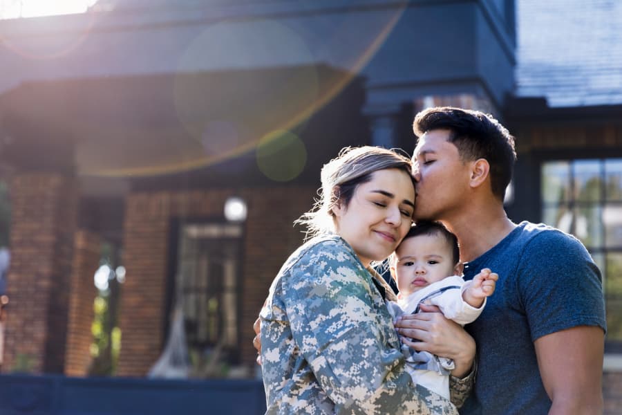 A man kisses his soldier wife, who is holding their baby